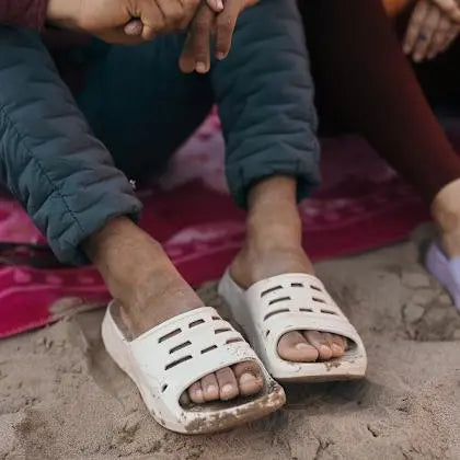 White sandals worn on feet sitting in sand with a person wearing blue pants and a dark jacket.