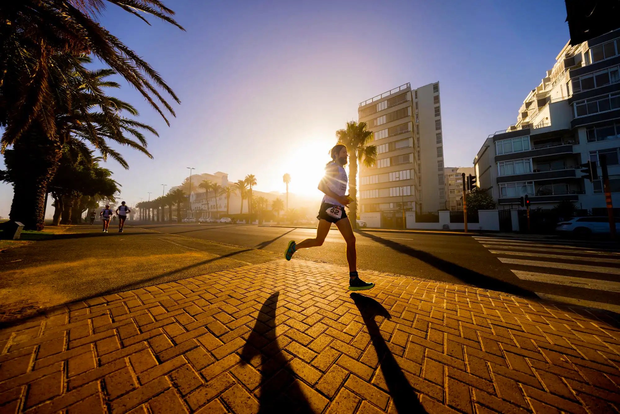 Person running on a brick path with palm trees and buildings in the background during sunset.