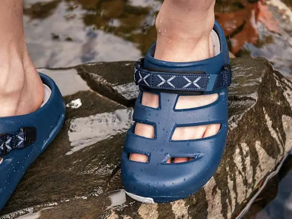 Navy blue sandals with white sole worn by a person standing on a wet surface.
