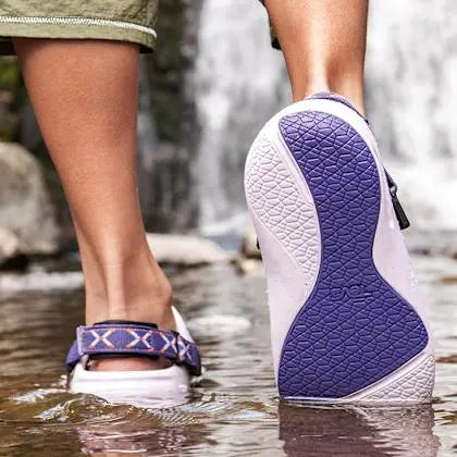 Person wearing purple and white wedge sandals walking through water with a blurred natural background