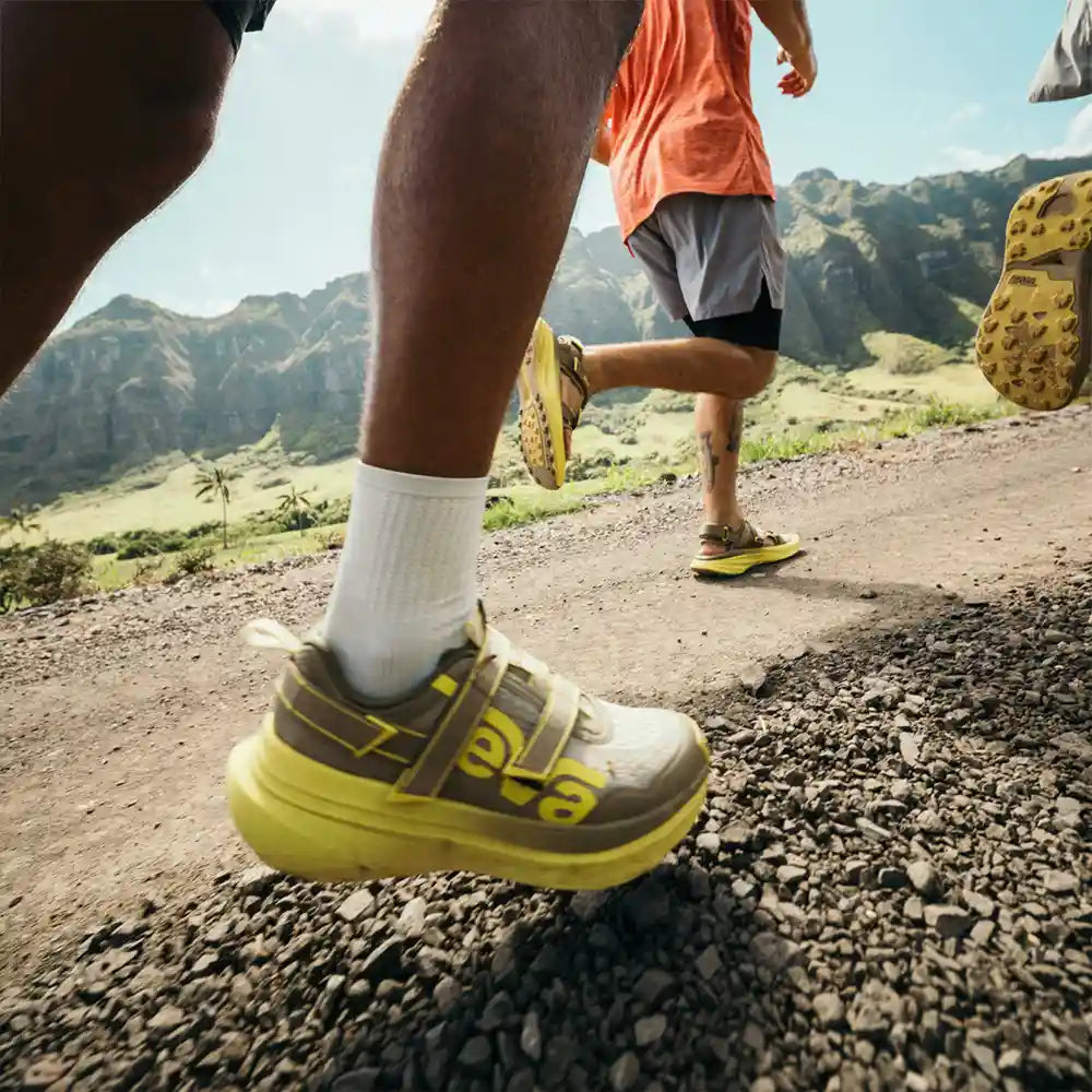 Two people running on a mountain trail wearing TEVA shoes.